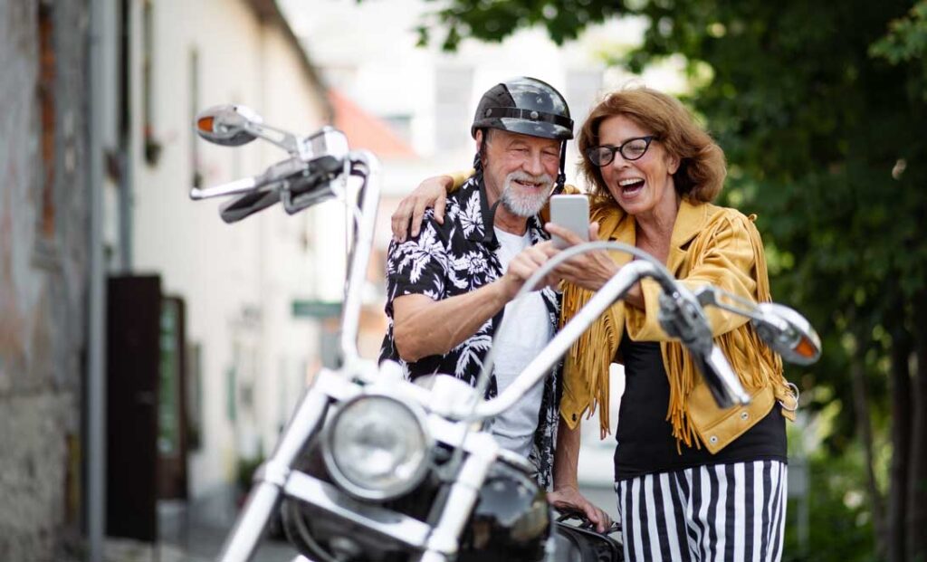 A front view of cheerful senior couple travellers with motorbike in town, taking selfie.